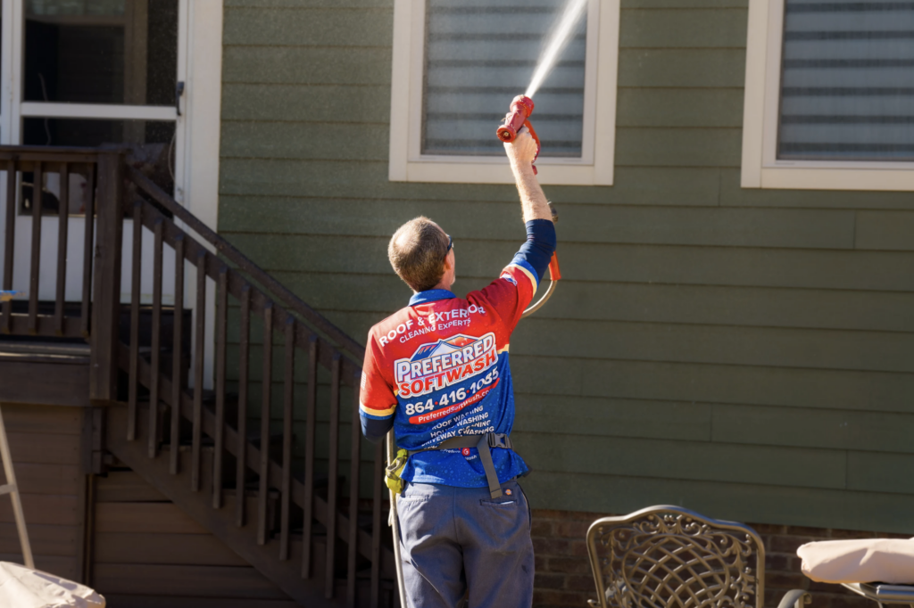 A person in a brightly colored uniform is power washing the exterior of a house. They are aiming the water spray at a closed window on the green siding. Nearby, a staircase and a patio table with chairs are visible.